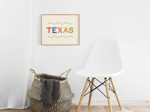 A modern white room with a white chair, a woven basket holding a gray blanket, and the Funky Texas Art Print by Onderkast Studio—bold colors and wavy green lines—framed on the wall.