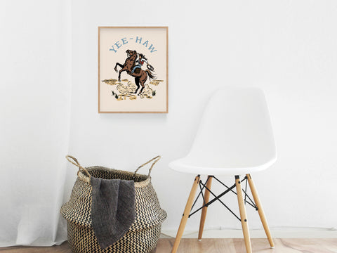 The Yeehaw Cowgirl Art Print by Onderkast Studio, featuring a bucking horse, is framed and displayed above a woven basket and modern white chair, adding country flair to this minimalist room.