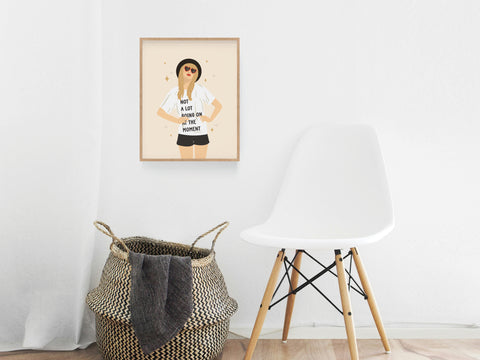 A minimal room with a white chair, woven basket, and the TS Red Art Print by Onderkast Studio—featuring a woman in sunglasses and a “Not a lot going on at the moment” shirt—hanging on the wall.