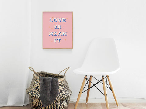 A white chair and woven basket sit in a minimalist room; the colorful Love Ya Mean It Art Print by Onderkast Studio hangs on the wall, adding a premium touch to the serene space.