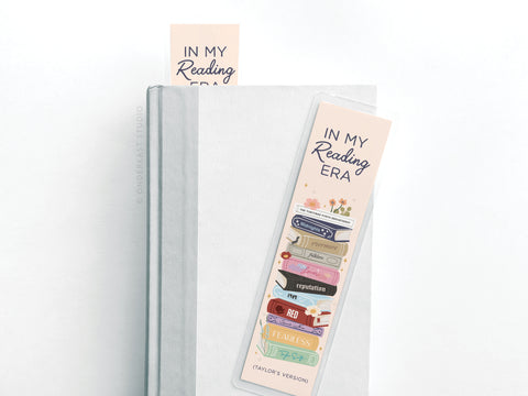 A white hardcover book displays the Easyhand Goods TS In My Reading Era Laminated Bookmark, featuring vibrant print with IN MY Reading ERA and a stack of colorful books named after Taylor Swift albums; another matching bookmark peeks out.