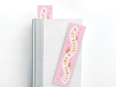 A light gray hardcover book features the Easyhand Goods Plot Twists & Hot Trysts Laminated Bookmark with playful pink lettering. Another durable bookmark from the same brand is partially visible at the top of the book.