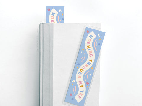 A closed hardcover book with Easyhand Goods Working Through My TBR Laminated Bookmark—featuring colorful text and fun shapes—partially inside, plus a matching sticker nearby for book lovers.