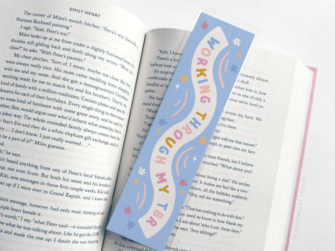 The Easyhand Goods Working Through My TBR Laminated Bookmark features colorful lettering and a playful blue design, making it a perfect gift for book lovers. It’s shown resting on an open book.
