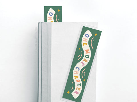 The Easyhand Goods One More Chapter Laminated Bookmark features colorful lettering on green and is ideal for avid readers, with one bookmark peeking from closed pages and another resting flat atop a white book.