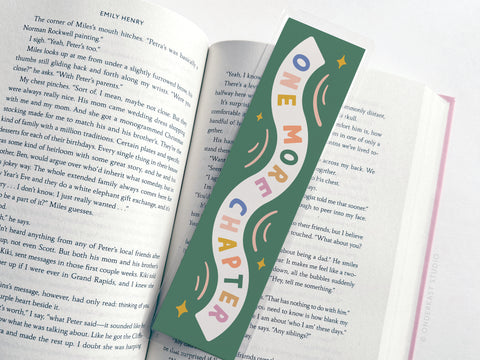 An open book features the Easyhand Goods “One More Chapter” Laminated Bookmark—a wavy, green design with rainbow and star art and playful lettering—making it a perfect gift for avid readers.