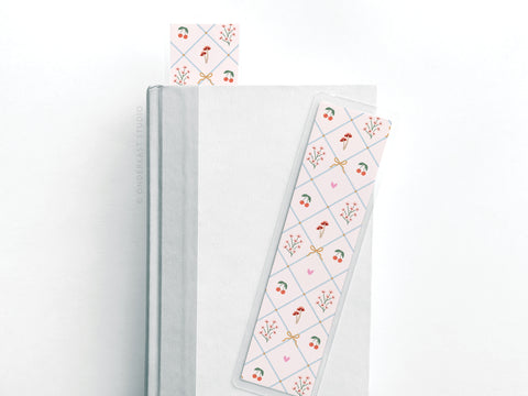 A white book with two Easyhand Goods Coquette Pattern Laminated Bookmarks—one inside, one on the cover. The bookmarks have pastel coquette designs with winter berries, holly, and hearts, and are printed in the USA.