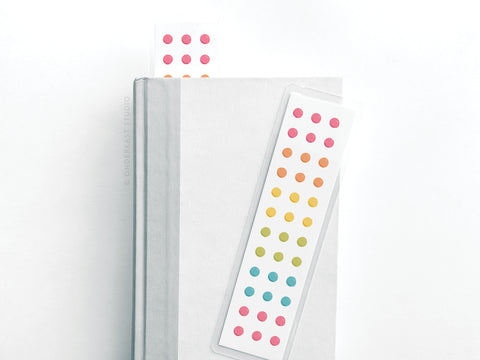 A white hardcover book with two Easyhand Goods Button Candy Laminated Bookmarks, featuring vibrant pink, orange, yellow, green, and blue dots, rests on a white surface.