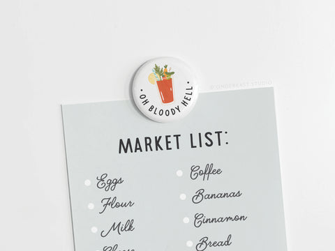 A grocery list clipped with the Onderkast Studio Oh Bloody Hell Bloody Mary Button Magnet—a round fridge magnet with a cocktail design and the phrase OH BLOODY HELL—lists eggs, flour, milk, coffee, bananas, cinnamon, and bread.
