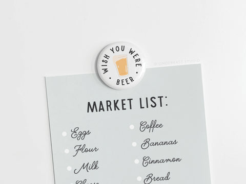 A grocery list with eggs, flour, milk, coffee, bananas, cinnamon, and bread is held by the Onderkast Studio Wish You Were Beer Button Magnet—featuring a beer glass and the phrase “Wish you were beer”—ideal for beer lovers or as a nostalgia gift.