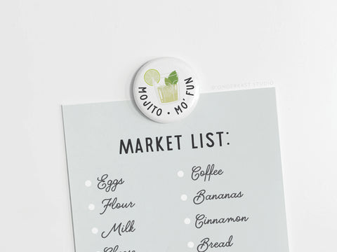 A shopping list is held by Onderkast Studio’s Mojito Mo’ Fun Button Magnet, featuring a mojito illustration and the phrase “Mojito Mo Fun.” The list includes eggs, flour, milk, cheese, coffee, bananas, cinnamon, and bread.