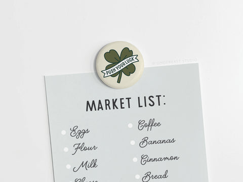 A grocery list featuring eggs, flour, milk, and coffee is held up by the Push Your Luck Clover Button Magnet from Onderkast Studio—a green four-leaf clover with a PUSH YOUR LUCK banner.