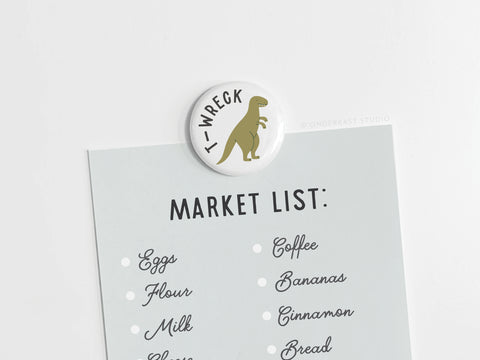 A grocery list is held on a white surface by the discontinued Onderkast Studio T-wreck Button Magnet, which features a colorful cartoon T. rex with T-WRECK text. The list includes eggs, flour, milk, coffee, bananas, cinnamon, and bread.
