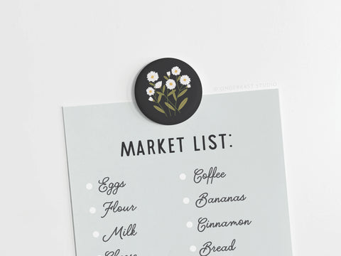 A grocery list is clipped with the Onderkast Studio Daisies Button Magnet, showcasing vibrant colors. Written in black cursive on a light gray sheet are eggs, flour, milk, coffee, bananas, cinnamon, and bread.