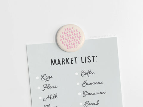 A grocery list on light gray paper is held by the discontinued YASS Pink Button Magnet from Onderkast Studio, featuring vibrant pink text reading YAAAAAAASSSS. The list includes eggs, flour, milk, cheese, coffee, bananas, cinnamon, and bread.