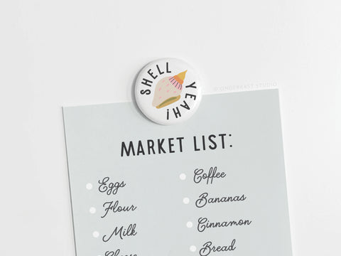 The Shell Yeah Button Magnet by Onderkast Studio features an illustrated seashell and text—perfect for keeping your essentials list, like eggs or coffee, organized for any beach lover or ocean enthusiast.