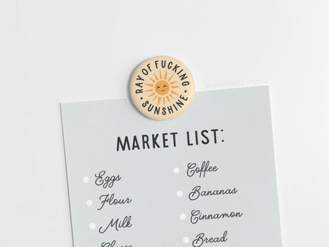 A grocery list is held up by the vibrant Ray of Fucking Sunshine Button Magnet from Onderkast Studio, featuring a smiling sun and bold lettering for a cheerful touch.