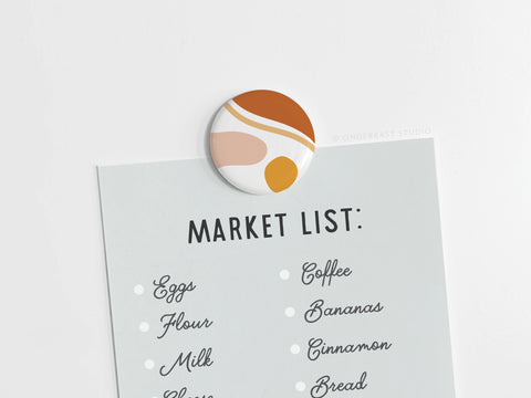 The Abstract No. 5 Button Magnet by Onderkast Studio holds a light-blue paper displaying “MARKET LIST:” and groceries like eggs, flour, milk, coffee, bananas, cinnamon, and bread. (This item has been discontinued.).