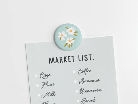 The Onderkast Studio White Flowers Button Magnet holds a light blue handwritten shopping list with eggs, milk, and bananas on a white surface.