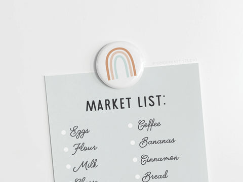 A light blue Market List is attached to a surface with the Rainbow Button Magnet by Onderkast Studio, which features a simple muted rainbow design. The list includes items such as eggs, flour, coffee, and bananas.