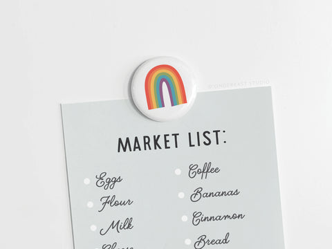 A grocery list is held to a surface with the Onderkast Studio Pride Rainbow Button Magnet, which features a vibrant rainbow flag design. The list shows eggs, flour, milk, coffee, bananas, cinnamon, and bread.