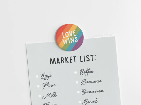 A grocery list with eggs, flour, milk, coffee, bananas, and bread is held up by the onderkast studio Love Wins Button Magnet, featuring rainbow stripes and LOVE WINS to celebrate LGBTQ+ pride.