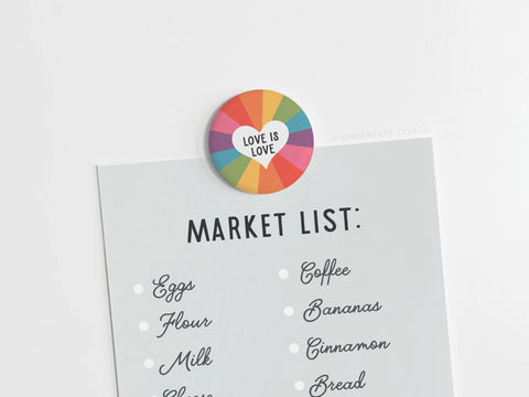 A Market List grocery list is held by the onderkast studio Love Is Love Button Magnet, which features vibrant colors and a heart design symbolizing love. Items on the list include eggs, flour, milk, coffee, bananas, cinnamon, and bread.