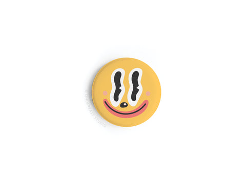 The Trippy Happy Face Button Magnet by Onderkast Studio is a round yellow magnet with wavy black eyes, a pink blush, and a wide red smile on a white background.