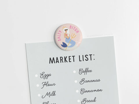 A market shopping list is held by the Salty Bitch Mermaid Button Magnet from Onderkast Studio, featuring a cartoon mermaid with a trident and “SALTY BITCH” text—ideal for beach lovers! List includes eggs, flour, milk, coffee, and bananas.