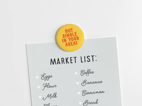 A shopping list with eggs, flour, milk, coffee, bananas, cinnamon, and bread is clipped by Onderkast Studios “Hot Single in Your Area!” button magnet—a humorous gift printed in the USA.