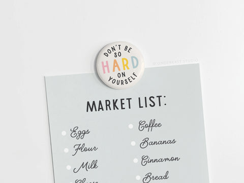 A light blue shopping list featuring groceries is held by Onderkast Studio’s “Don’t Be So Hard On Yourself” Button Magnet, promoting self-compassion. Made in the USA.
