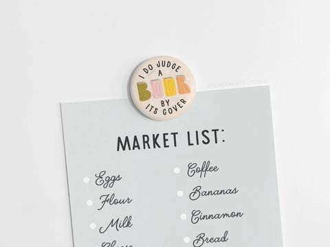 The Onderkast Studio I DO Judge a Book By Its Cover button magnet playfully secures a shopping list—eggs, flour, milk, coffee, bananas, cinnamon, and bread—on a light surface with its vibrant round design.