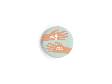 The Hug Me Hands Button Magnet by Onderkast Studio features two illustrated hands reaching toward each other with hug me between them on a pale blue background—a playful invitation to connect.