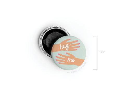 The Hug Me Hands Button Magnet by Onderkast Studio features two illustrated arms reaching out with playful white text. This 1.25-inch accessory has an open black backing and is also available as a classic pinback button.