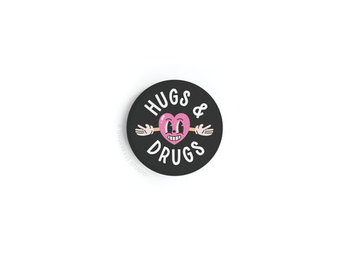 The Hugs & Drugs Button Magnet by Onderkast Studio is a round black magnet with “HUGS & DRUGS” in white, featuring a smiling pink cartoon heart with outstretched arms, celebrating the joy of physical touch.