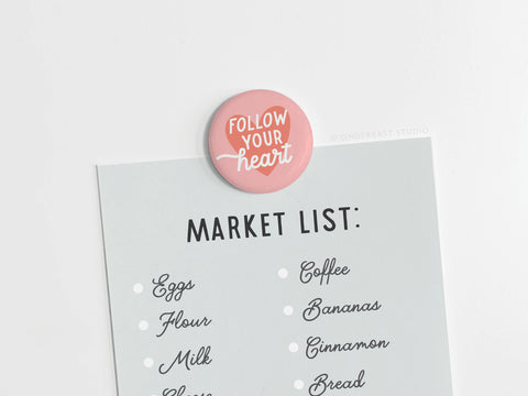 A paper market list with eggs, flour, milk, coffee, and bananas is held by a vibrant Follow Your Heart Deep Button Magnet from Onderkast Studio in pink with white script.