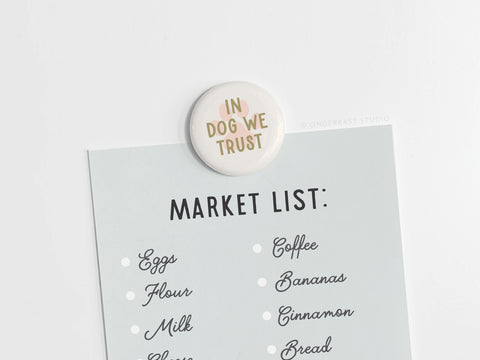 The Onderkast Studio In Dog We Trust Button Magnet holds a light blue market list with groceries like eggs, flour, milk, coffee, bananas, cinnamon, and bread on a white surface—ideal for dog-loving households.
