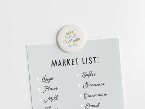 The Onderkast Studio Great Minds Overthink Alike Button Magnet humorously holds a shopping list—with eggs, flour, milk, cheese, coffee, bananas, cinnamon, and bread—on a white surface.