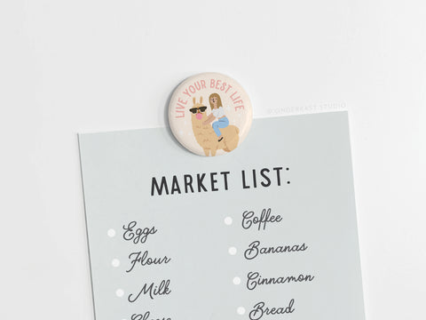 The Onderkast Studio Live Your Best Life Button Magnet displays a colorful cartoon llama and holds a shopping list with eggs, flour, milk, coffee, bananas, and bread on a white surface.
