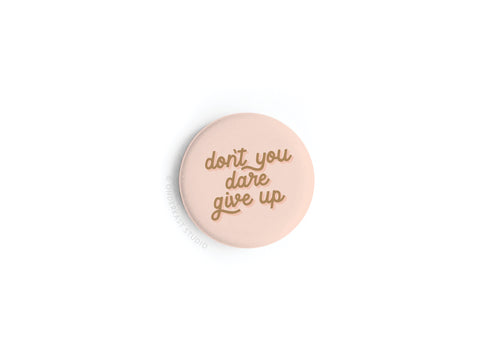 The Don’t You Dare Give Up Button Magnet by Onderkast Studio features gold script on a round, pale pink design—an inspiring, USA-printed motivational magnet set against a white background.