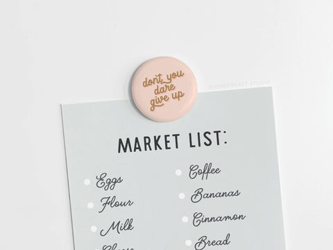 A shopping list is held by the Onderkast Studio “Don’t You Dare Give Up Button Magnet,” a round, pale pink motivational magnet made in the USA. The handwritten list includes eggs, flour, milk, coffee, bananas, cinnamon, and bread.
