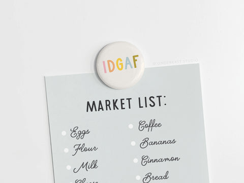 A light blue grocery list is held to a white surface by the IDGAF Button Magnet from Onderkast Studio, adding a playful and confident accent to your kitchen routine.