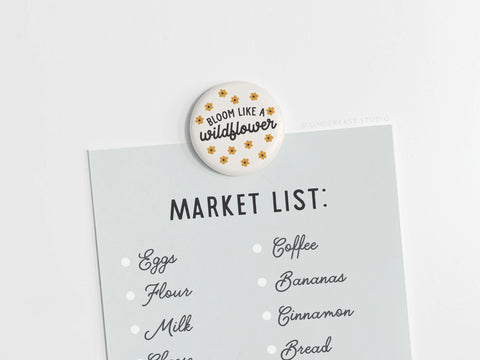 Onderkast Studio’s Bloom Like a Wildflower Button Magnet, featuring yellow flowers and uplifting text, secures a market list—eggs, flour, milk, coffee, bananas, cinnamon, bread—on a white surface.