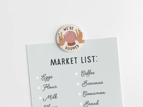 The We’re Doomed Button Magnet by Onderkast Studio, featuring vivid colors and illustrated hands with a crystal ball, holds your market list on any light surface. Proudly printed in the USA.