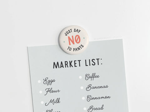The Onderkast Studio Just Say No to Pants Button Magnet holds a market list—eggs, flour, milk, coffee, bananas, cinnamon, and bread—on a light surface, celebrating comfort and a pants-free lifestyle.
