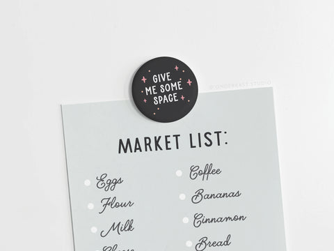 A grocery list is held by the Onderkast Studio Give Me Some Space Button Magnet, reminding everyone of personal space. On the list: eggs, flour, milk, coffee, bananas, cinnamon, and bread.