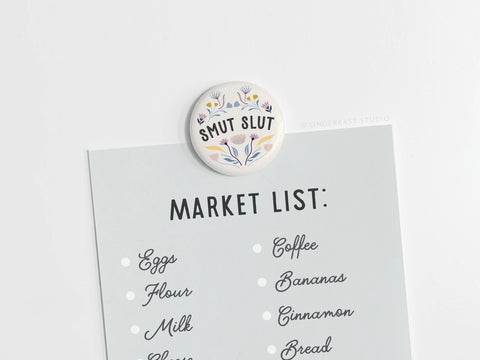 A grocery list on light blue paper is held by an Onderkast Studio Smut Slut Button Magnet, which has a floral motif and playful literary humor. The list features eggs, flour, coffee, and bananas.