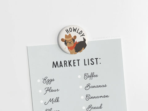 The Howldy Button Magnet by Onderkast Studio is a Western-inspired accessory featuring a cartoon dog in a cowboy hat and red bandana, with vibrant colors—perfect for holding your market list of eggs, flour, milk, coffee, bananas, cinnamon, and bread.