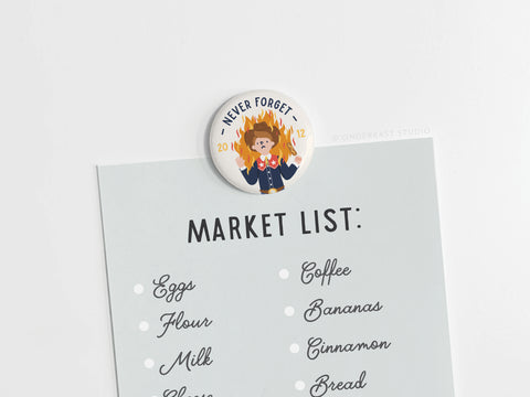 The Onderkast Studio Never Forget Button Magnet, featuring vibrant colors and a dog amid flames, adds regional humor while holding notes—like eggs or coffee—securely to any surface.