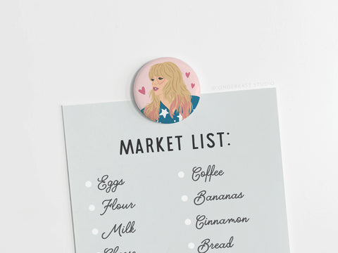 A grocery list is held on the fridge by the Onderkast Studio TS Lover Button Magnet, a cute kitchen accent featuring an illustrated blonde woman with bangs and pink hearts. The list includes eggs, flour, milk, coffee, bananas, cinnamon, and bread.